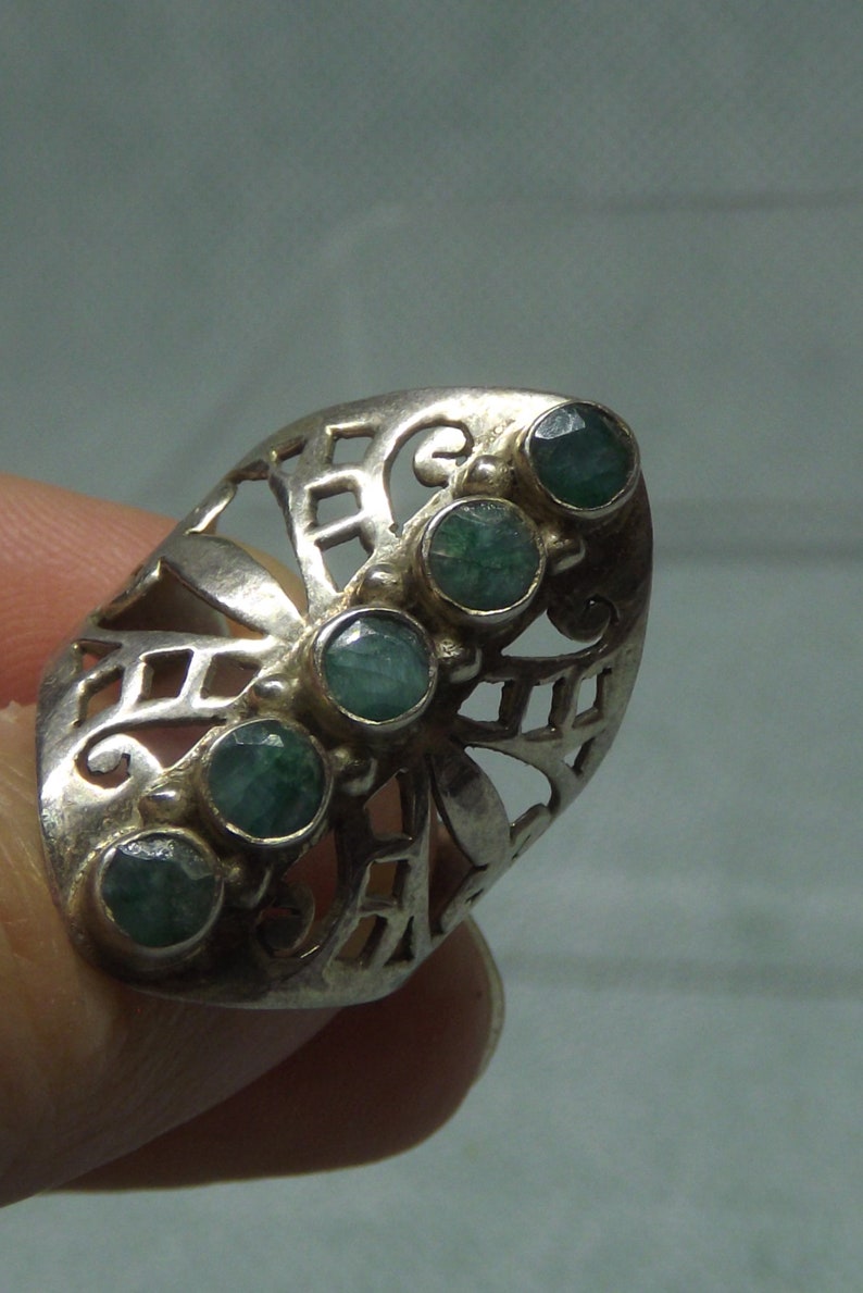 May include: A silver ring with a filigree design and five green gemstones. The ring is on a finger.