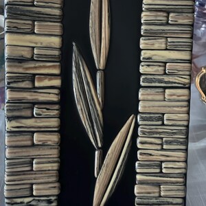 May include: A decorative wall art piece featuring a black background with a central design of three stylized leaves in shades of beige and brown. The sides are bordered with rectangular blocks in similar tones, creating a textured effect.
