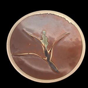 May include: A round ceramic plate with a brown center and a cream-colored rim. The plate features a stylized plant design in shades of brown, black, and green. The design is centered on the plate, with the plant's leaves extending outwards.