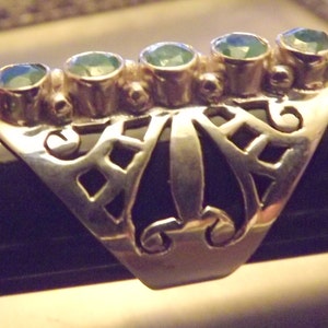 May include: A silver ring with a filigree design and five green gemstones. The ring is set on a black surface.