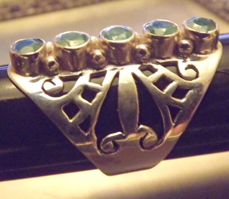 May include: A silver ring with a filigree design and five green gemstones. The ring is set on a black surface.