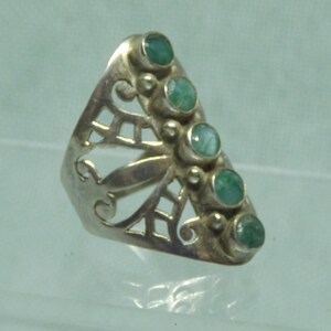 May include: A silver ring with a delicate, openwork design and six green gemstones. The ring is set on a light blue background.