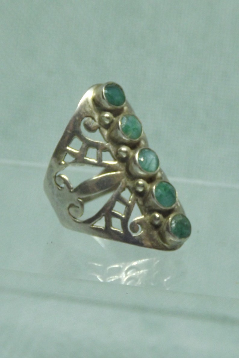 May include: A silver ring with a delicate, openwork design and six green gemstones. The ring is set on a light blue background.