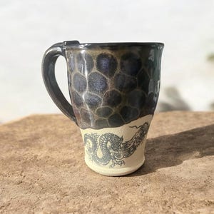 May include: A ceramic mug with a black handle and a dark, textured upper half. The lower half is cream-colored with a black dragon design. The mug is sitting on a brown surface.