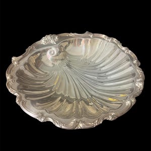 May include: A silver-colored, oval-shaped serving dish with a shell-like design. The dish has a scalloped edge with ornate detailing. The interior has a reflective surface, and the background is black.