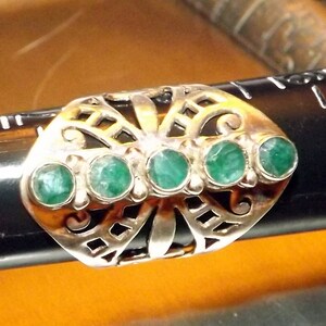 May include: A silver ring with a butterfly design and five green gemstones. The ring is set on a black surface.