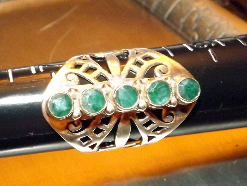 May include: A silver ring with a butterfly design and five green gemstones. The ring is set on a black surface.