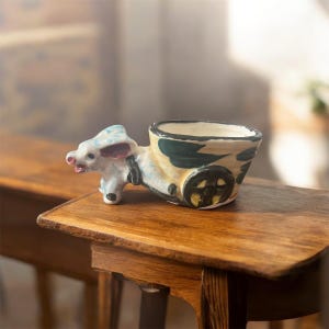 May include: A ceramic planter shaped like a cow pulling a cart. The cow is white with blue and pink accents, and the cart is beige with green and black details. The planter has a small opening for plants.