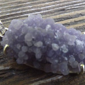 Puede incluir: Un colgante de color lavanda con un grupo de pequeñas formaciones redondeadas. El colgante está engastado en metal plateado con detalles ornamentados en la parte superior. La piedra tiene un aspecto texturizado y el engaste incluye un pequeño lazo dorado.