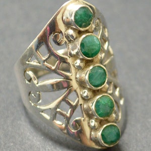 May include: A silver ring with a delicate, openwork design and five emerald green gemstones set in a row.