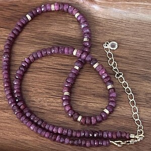 Könnte beinhalten: Eine Halskette aus kleinen, facettierten, violetten Edelsteinen mit goldenen Akzenten. Die Halskette liegt auf einer Holzoberfläche.