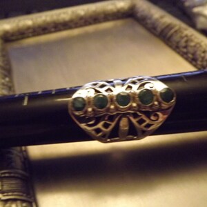 May include: A silver ring with a filigree design and five green gemstones. The ring is set on a black surface.
