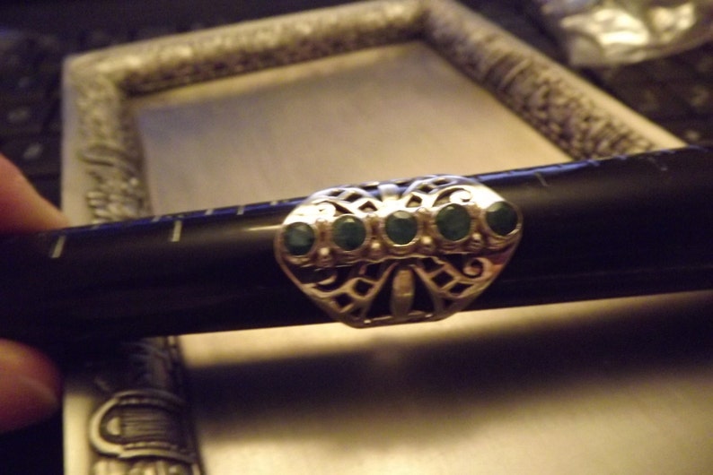 May include: A silver ring with a filigree design and five green gemstones. The ring is set on a black surface.