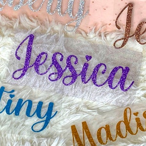 May include: Five glitter-covered names, "Destiny", "Jessica", "Madison", "Jess", and "Emily", are displayed on a white fluffy surface. The names are cut from vinyl and have a sparkly finish.
