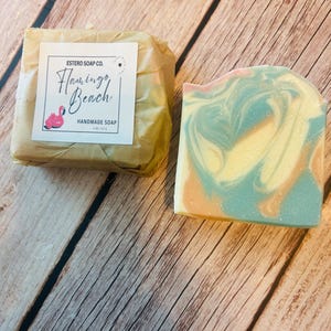 Pode incluir: Duas barras de sabonete artesanal em uma superfície de madeira. Uma barra é embrulhada em papel pardo com um rótulo que diz "Flamingo Beach Handmade Soap". A barra desembrulhada tem um design em espiral em tons de azul, amarelo e pêssego.