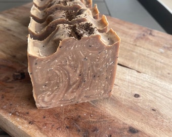 Op de afbeelding: Een rechthoekige blok handgemaakte zeep met een lichtbruine kleur en een textuur. De zeep heeft een wervelpatroon en is bedekt met donkerbruine korrels. De zeep ligt op een houten snijplank.