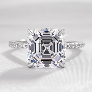 May include: A square-cut diamond engagement ring with a pavé band. The large, clear center stone is set in a four-prong setting. The band is made of silver-colored metal and is lined with small, sparkling stones. The ring is displayed on a white, textured surface.