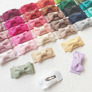Könnte beinhalten: Eine Sammlung von Haarspangen mit kleinen Schleifen aus geripptem Stoff in einer Vielzahl von Farben, darunter Rosa, Rot, Braun, Grün, Blau, Gelb und Weiß. Jede Schleife ist an einer Metallspange befestigt, die auf der Unterseite sichtbar ist.