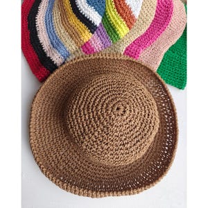May include: A brown, crocheted sun hat with a wide brim, displayed with a variety of colorful crocheted hats in the background. The hats are in shades of red, black, blue, white, yellow, orange, pink, and green. The hats are handmade.