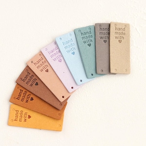 May include: A set of eight leather tags in various colours, each with the text "hand made with" and a heart symbol. The tags are arranged in a semi-circle.