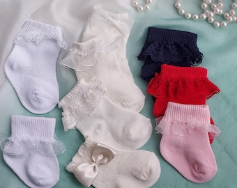 Könnte beinhalten: Eine Auswahl an Babysocken in Weiß, Marineblau, Rot und Rosa. Einige Socken haben Spitzensaum, andere Satin-Schleifen. Die Socken sind auf einer hellblauen Oberfläche mit einer Perlenkette im Hintergrund angeordnet.