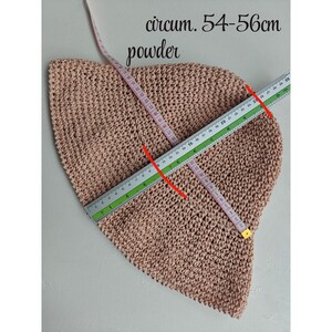 May include: A powder-colored, crocheted hat with a circumference of 54-56cm. The hat is measured with a ruler and a measuring tape. The text "circum. 54-56cm" and "powder" are visible in the image. The hat is likely handmade.