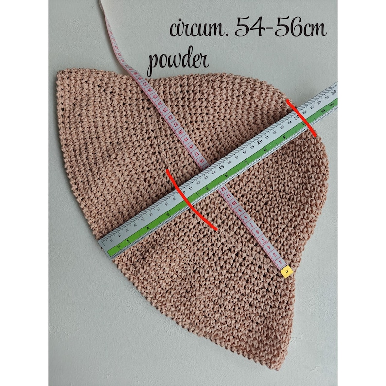 May include: A powder-colored, crocheted hat with a circumference of 54-56cm. The hat is measured with a ruler and a measuring tape. The text "circum. 54-56cm" and "powder" are visible in the image. The hat is likely handmade.