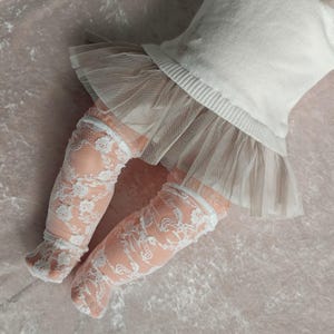 May include: A white sweater with a tulle skirt and peach-colored lace socks. The socks are adorned with a delicate white floral lace pattern. The outfit is set against a soft, pink background.