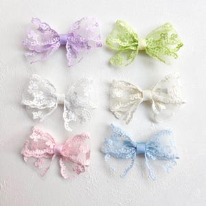 Könnte beinhalten: Sechs Haarschleifen aus Spitze in Pastellfarben: Lavendel, Grün, Weiß, Elfenbein, Rosa und Blau. Jede Schleife hat ein zartes florales Spitzendesign und eine passende Mittelband. Die Schleifen sind auf einer weißen, strukturierten Oberfläche angeordnet.