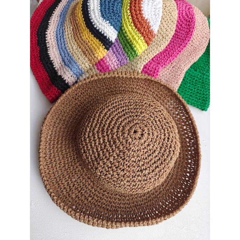 May include: A brown, crocheted sun hat with a wide brim, displayed with a variety of colorful crocheted hats in the background. The hats are in shades of red, black, blue, white, yellow, orange, pink, and green. The hats are handmade.