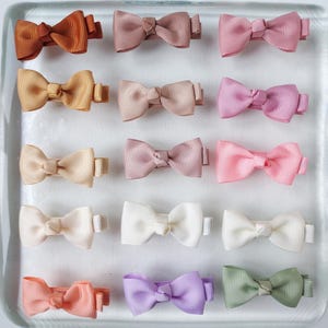 Pode incluir: Uma coleção de doze pequenos laços de cabelo de fita em várias cores, incluindo tons de castanho, rosa, pêssego, lavanda e verde. Cada laço é preso a uma pequena presilha de metal. Os laços estão dispostos em um padrão de grade.