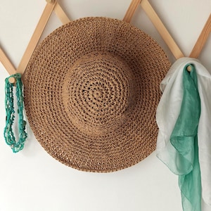 May include: A natural-toned woven sun hat with a wide brim hangs on a wooden rack. A turquoise beaded necklace and a white and teal scarf are also displayed. The hat's design suggests a focus on sun protection.