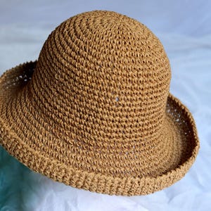 May include: A woven, tan-colored sun hat with a rounded crown and a wide, downward-sloping brim. The hat is made of a textured material, likely straw or a similar natural fiber. The brim measures approximately 4 inches wide.