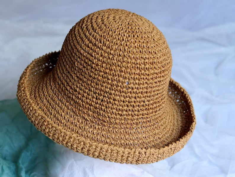 May include: A woven, tan-colored sun hat with a rounded crown and a wide, downward-sloping brim. The hat is made of a textured material, likely straw or a similar natural fiber. The brim measures approximately 4 inches wide.
