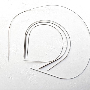 May include: Several silver knitting needles of different sizes are displayed on a textured white surface. The needles are curved and pointed, designed for circular knitting. The image highlights the tools used in the craft of knitting.