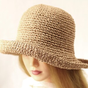 May include: A tan, crocheted bucket hat with a slightly curved brim. The hat is made of a woven material and has a rounded crown. The hat is shown on a mannequin head with blonde hair.