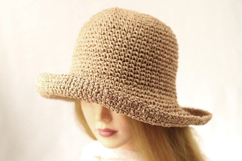 May include: A tan, crocheted bucket hat with a slightly curved brim. The hat is made of a woven material and has a rounded crown. The hat is shown on a mannequin head with blonde hair.
