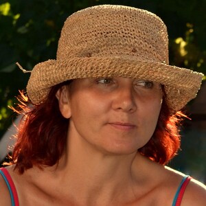 May include: A woven straw hat with a wide brim. The hat is a light tan color and has a slightly rounded crown. The person is wearing a red and blue top.