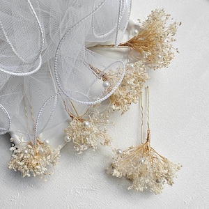 Op de afbeelding: Een verzameling bruids haaraccessoires, waaronder haarspelden en een sluier, gerangschikt op een wit oppervlak. De haarspelden zijn voorzien van gedroogde bloemen en imitatieparels. De sluier is gemaakt van witte tule met zilveren rand.