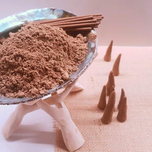 以下が含まれることがあります： アワビの貝殻のボウルには茶色の粉末とお香スティックが入っており、白い木製のスタンドに置かれています。茶色の香りのコーンがいくつか近くに配置されています。ボウルは真珠のような虹色の光沢があります。