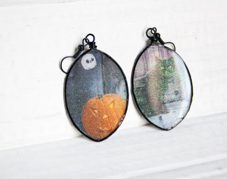 May include: A pair of Halloween-themed earrings with black wire frames. One earring features a glittery image of a jack-o'-lantern, while the other features a glittery image of a black cat.