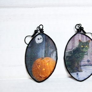 May include: A pair of Halloween-themed earrings. One earring features a black cat with green eyes, and the other earring features a jack-o'-lantern with a spooky face.