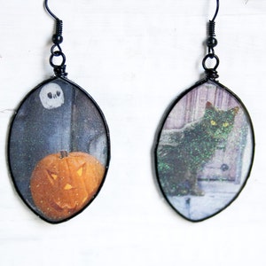 May include: A pair of black wire earrings with oval-shaped resin charms. One charm features a black cat with green eyes and the other charm features a jack-o'-lantern.