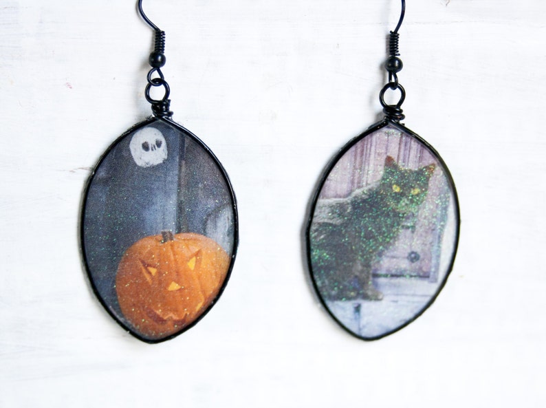 May include: A pair of black wire earrings with oval-shaped resin charms. One charm features a black cat with green eyes and the other charm features a jack-o'-lantern.