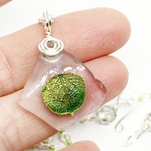 May include: A silver chain necklace with a clear resin pendant featuring a preserved green leaf. The pendant is shaped like a diamond and has a silver wire wrapped around the top.
