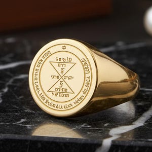 Puede incluir: Un anillo de sello dorado con un diseño circular grabado. El diseño incluye texto hebreo y formas geométricas. El anillo se muestra sobre una superficie de mármol blanco y negro. Una caja de madera y una perla están en el fondo.