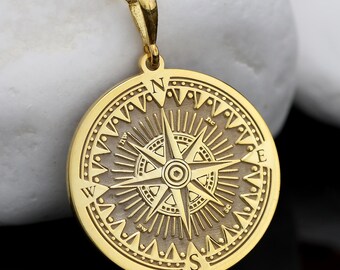 May include: A gold compass pendant necklace. The pendant has a detailed compass rose design with directional markers. The necklace is displayed on a white stone surface, with a dark background and orange berries.