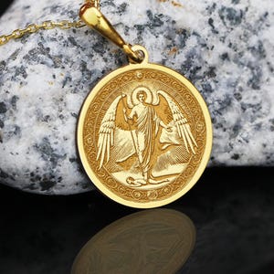 May include: A gold pendant necklace with a detailed engraving of an angel with outstretched wings. The round pendant hangs from a delicate gold chain. The angel is depicted in a flowing robe, holding a staff. The background is a textured stone.