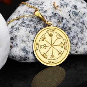 May include: A gold-coloured pendant necklace. The circular charm features Hebrew text and symbols. The pendant hangs from a fine gold chain. The necklace is displayed on a grey and white stone surface.