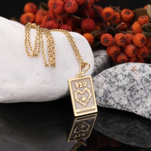 May include: Gold-colored pendant necklace with a rectangular charm. The charm features the number "420" above a heart-shaped design with a cannabis leaf. The words "THE STONER" are below the heart. The necklace is displayed on a white stone.